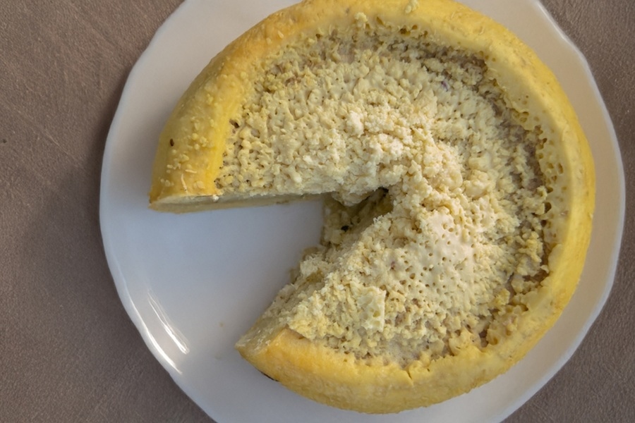 Un queso circular de color amarillo con textura grumosa reposa sobre un plato blanco al que le falta una porción triangular.