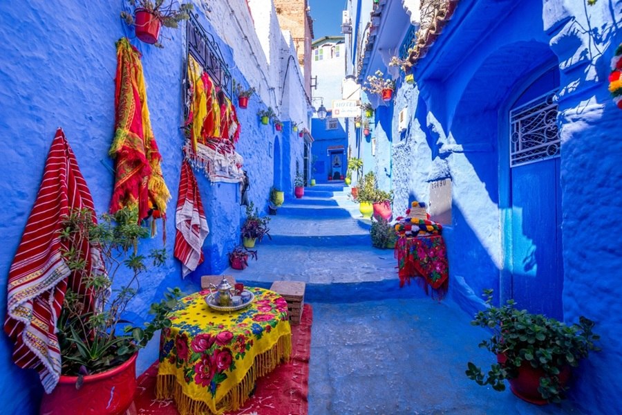 Una estrecha callejuela escalonada teñida de azul intenso se extiende entre fachadas decoradas con alfombras coloridas y macetas floridas, con una mesita de té tradicional en primer plano.