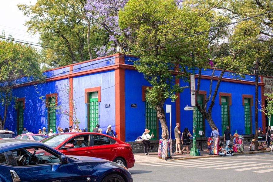 La fachada azul cobalto del Museo Frida Kahlo se alza tras peatones y tráfico en una calle arbolada. Texto visible: "MUSEO FRIDA KAHLO".