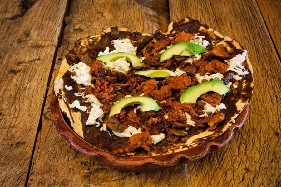Una tlayuda oaxaqueña reposa sobre un plato de barro artesanal. Muestra una base de tortilla tostada cubierta con frijoles, queso oaxaca deshebrado, carne preparada y rodajas de aguacate fresco sobre una mesa de madera.