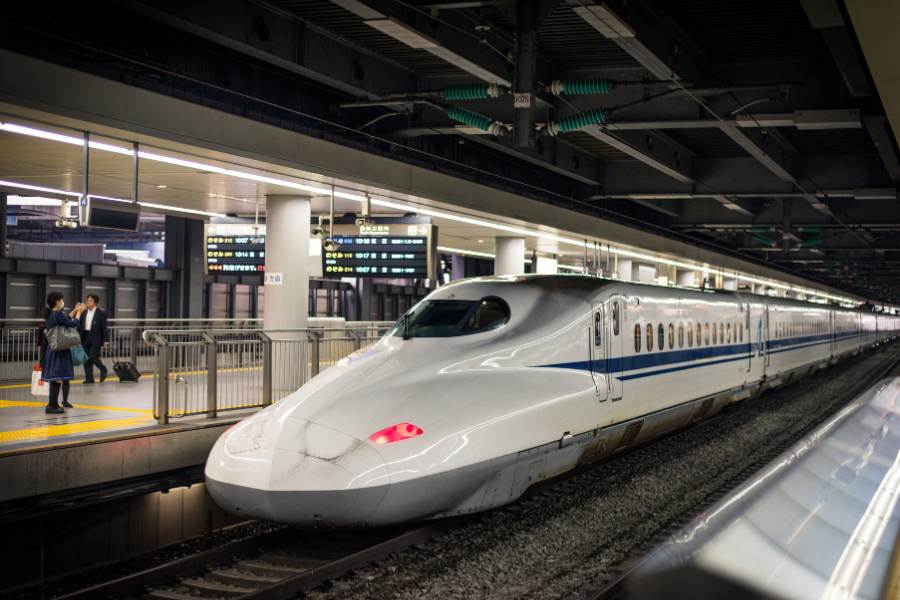 Un tren Shinkansen blanco de alta velocidad permanece estacionado en la vía de una moderna estación ferroviaria de Japón, Tokio, techada, mientras una pasajera toma una fotografía desde el andén iluminado.