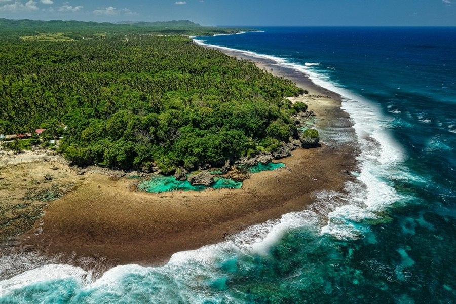 Una frondosa selva tropical bordea una costa rocosa donde olas blancas rompen suavemente contra piscinas naturales turquesas y el profundo océano azul.