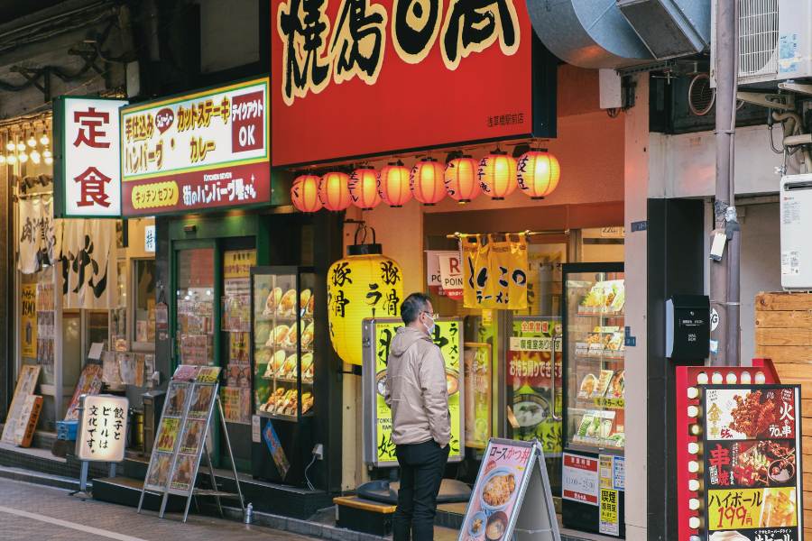 Un peatón de espaldas observa las vitrinas con réplicas de comida de un restaurante callejero tradicional iluminado por farolillos naranjas y carteles en una acera de Tokio.