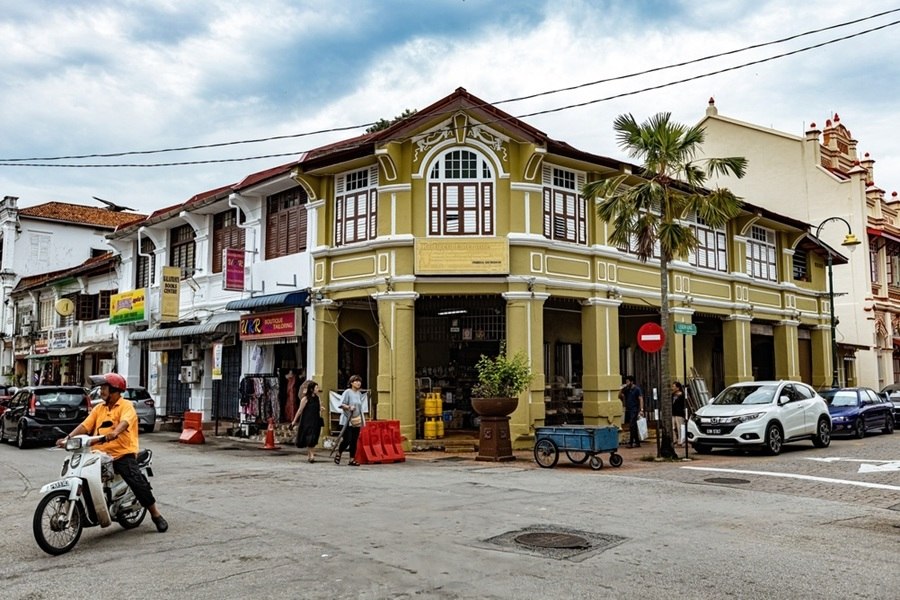Un motociclista circula frente a pintorescos edificios de estilo colonial en una esquina del casco histórico de George Town, entre transeúntes y coches aparcados bajo un cielo nublado.