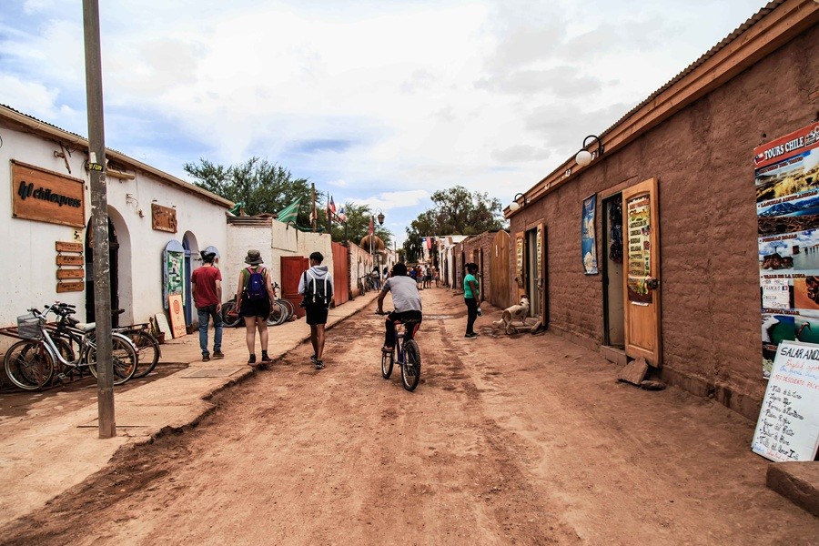 Varias personas y un ciclista recorren una calle de tierra flanqueada por edificios rústicos de adobe en un pueblo desertico. Texto en la imagen: "chilexpress", "TOURS CHILE", "SALAR ANDINO".