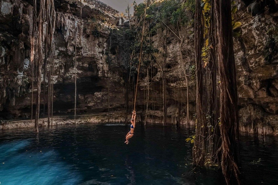 Una mujer en bikini se balancea colgada de una cuerda hacia el agua cristalina de un cenote natural rodeado de altas paredes rocosas y largas raíces colgantes.
