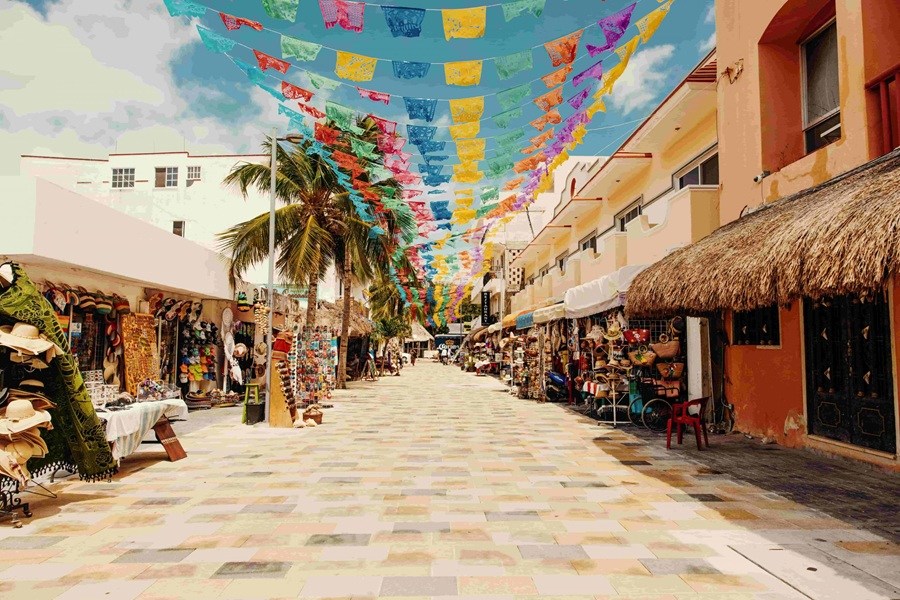 Cuelgan hileras de papel picado multicolor sobre una calle peatonal empedrada, flanqueada por tiendas de recuerdos localizadas entre palmeras y edificios coloridos bajo un cielo despejado.