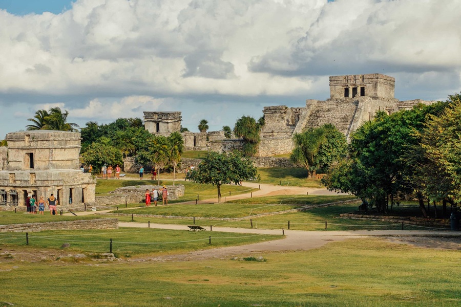 Ruinas mayas de piedra erigidas sobre un prado verde mientras varios turistas pasean por caminos de tierra rodeados de vegetación tropical bajo un cielo nublado.