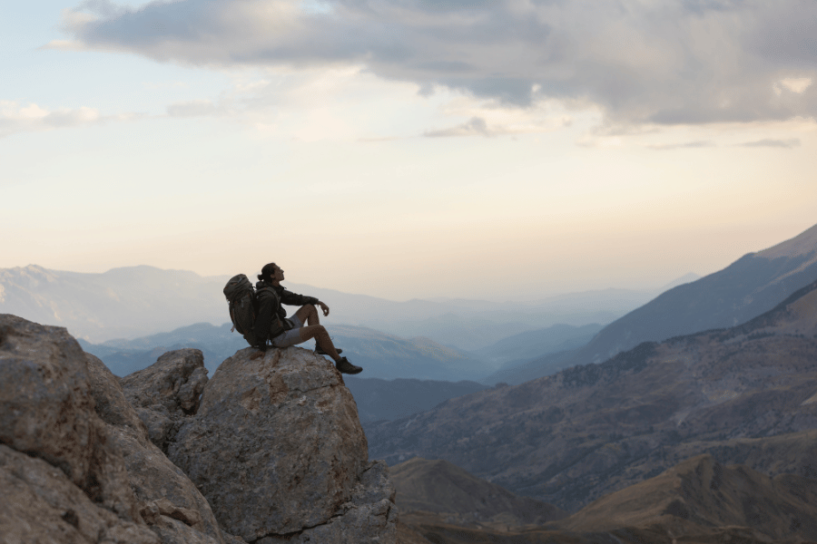 Una persona con mochila se sienta en una formación rocosa, mirando hacia un valle y montañas distantes bajo un cielo nublado.