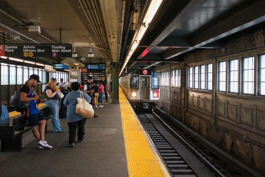 Un tren de metro entra en una estación cubierta mientras varios pasajeros esperan en el andén junto a carteles indicativos. T