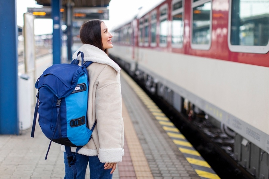 Una mujer sonriente con abrigo claro y mochila azul camina por el andén de una estación junto a un tren de pasajeros rojo y blanco.