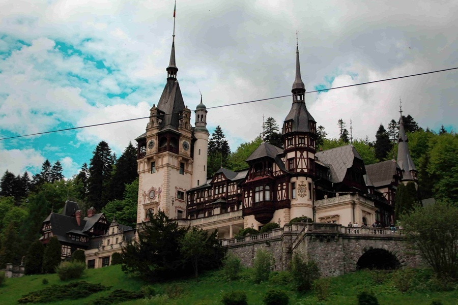 El Castillo de Peleș se alza imponente con sus torres puntiagudas y entramados de madera, rodeado de colinas verdes y densos bosques bajo un cielo nublado.