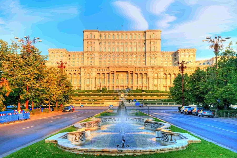 El Palacio del Parlamento de Bucarest se alza de fondo, mientras una fuente ornamental echa agua en primer plano en medio de una avenida arbolada con tráfico moderado bajo un cielo claro.