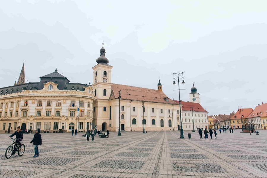 Varias personas pasean y montan en bicicleta por la gran plaza adintelada de Sibiu, rodeada por elegantes edificios históricos y torres de iglesia bajo un cielo encapotado.