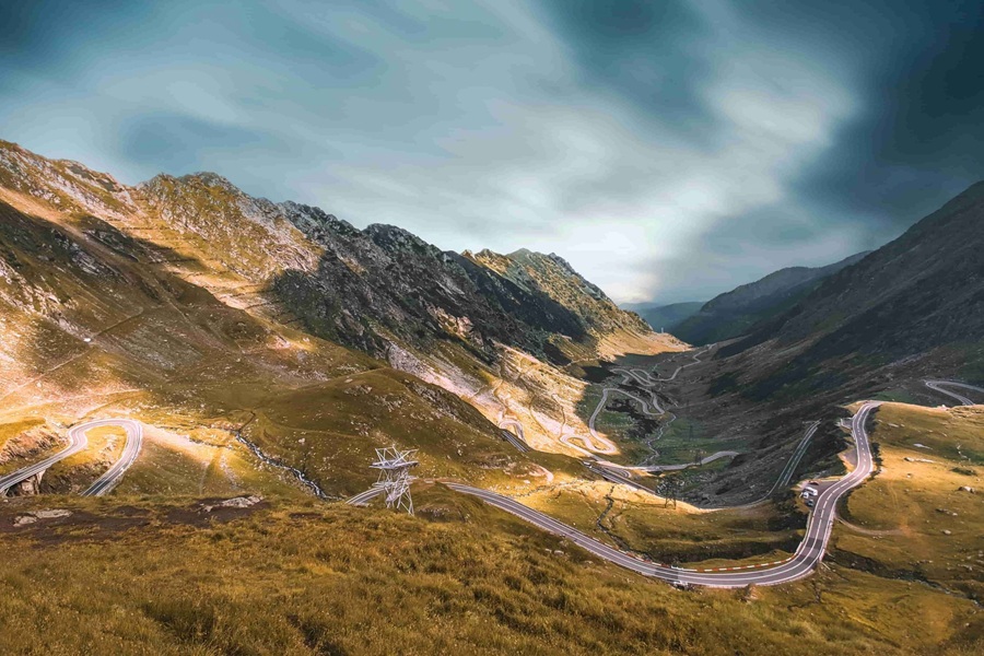 La carretera Transfăgărășan serpentea en curvas pronunciadas a través de un valle montañoso, rodeada de laderas rocosas y bajo un cielo dramático y nublado.
