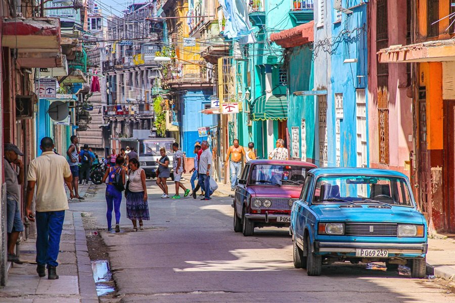 Varias personas caminan y conversan por una estrecha calle urbana flanqueada por edificios coloridos y coches antiguos aparcados a los lados. En las matrículas se lee: "P054" y "P069 249".