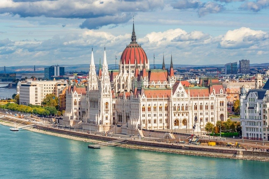 El Parlamento de Budapest destaca con sus agujas neogóticas a orillas del río Danubio, bajo un cielo parcialmente nublado con vistas al horizonte urbano de la ciudad.
