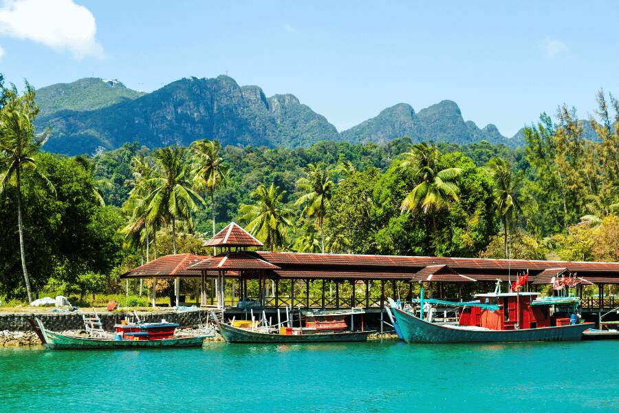 Barcos pesqueros de madera flotan en aguas turquesas junto a un embarcadero, con montañas selváticas al fondo en la isla de Langkawi, Malasia.