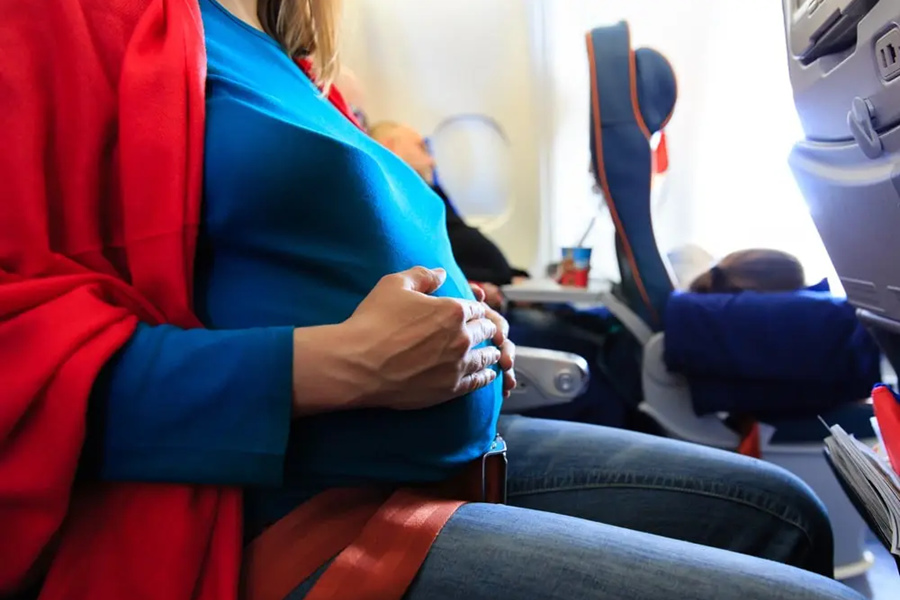 Una mujer embarazada con camiseta azul sostiene su vientre con ambas manos mientras viaja sentada con el cinturón de seguridad abrochado en el asiento de un avión de pasajeros.