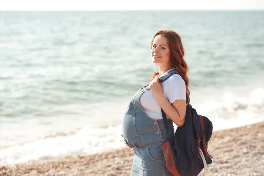 Una mujer embarazada con peto vaquero y mochila se sostiene la barriga mientras sonríe de pie en la orilla de una playa con el mar de fondo.