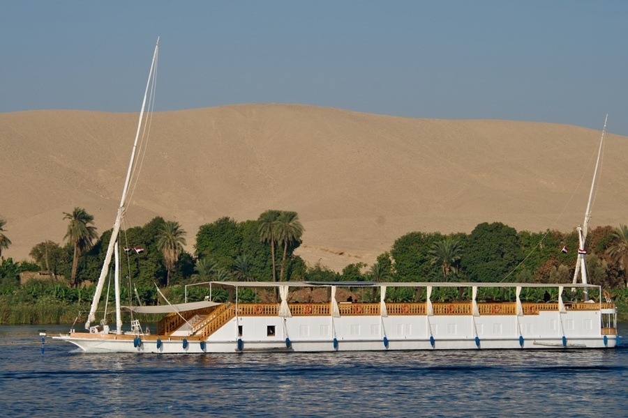 Una embarcación dahabiya blanca navega por las aguas del río Nilo, frente a una orilla con frondosas palmeras y una gran duna de arena dorada al fondo.