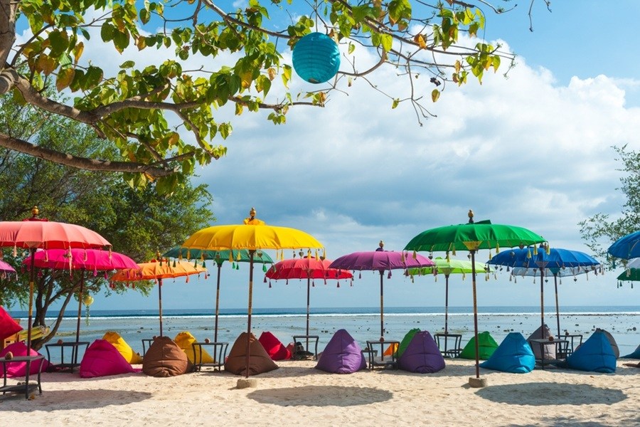Puffs de colores y sombrillas tradicionales proyectan sombra sobre la arena en una playa tropical, ofreciendo un espacio de descanso junto al mar bajo un cielo parcialmente nublado.