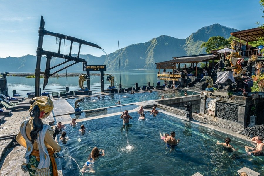 Varias personas se bañan en piscinas termales al aire libre, rodeadas de estatuas y fuentes de piedra, con un gran lago y montañas al fondo bajo un cielo despejado.