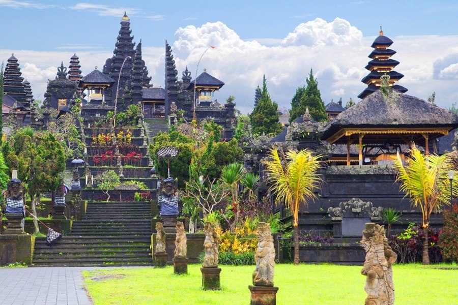 El templo de Pura Besakih se erige con sus torres de varios niveles sobre escalinatas de piedra y estatuas tradicionales, rodeado de vegetación tropical bajo un cielo nublado.