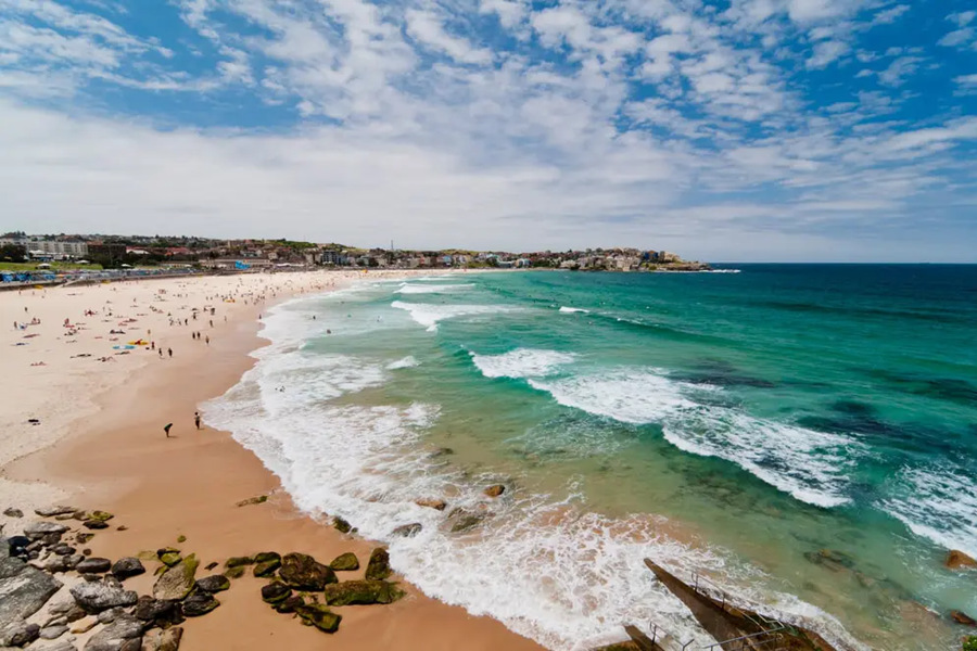 Las olas del océano turquesa rompen progresivamente en la orilla de una extensa playa de arena poblada de gente en Bondi Beach, Sídney, bajo un cielo despejado.