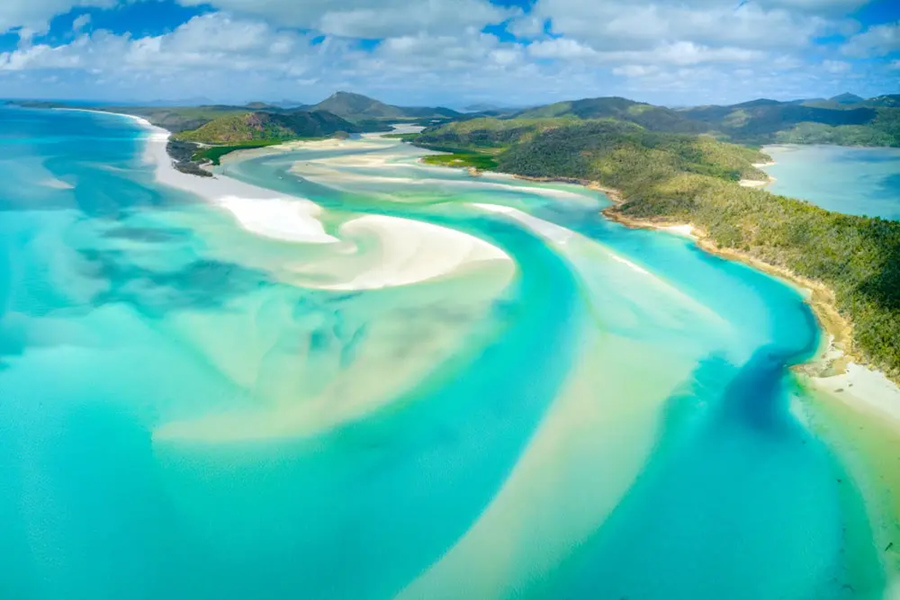 Aguas turquesas y bancos de arena blanca se entrelazan creando patrones sinuosos a lo largo de una costa tropical con colinas verdes bajo un cielo azul con nubes dispersas.