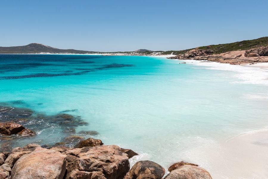 Parque Nacional Cabo Le Grand en Australia. Playa con agua turquesa y cristalina azul celeste, orilla con arena blanca y montañas al fondo con hierba verde oscura