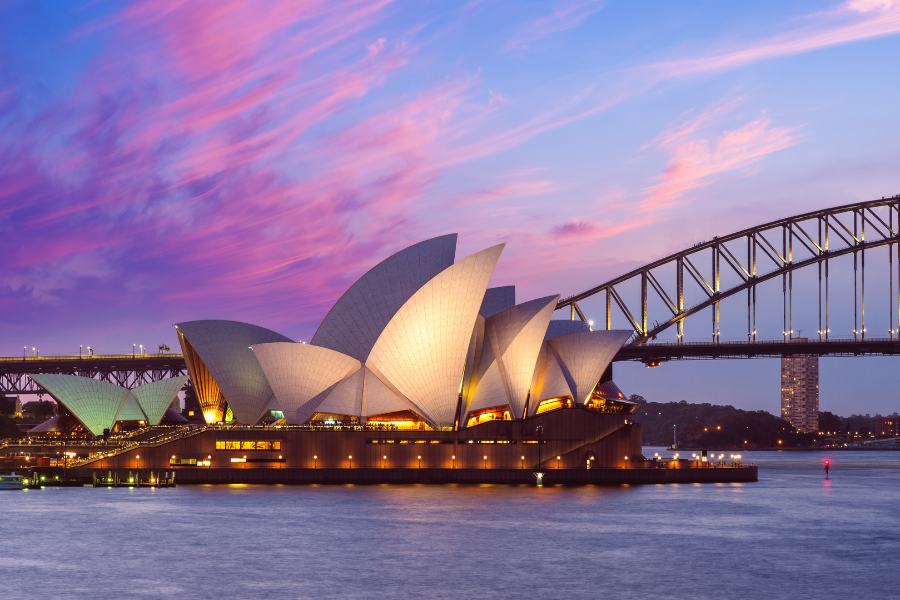 monumento más visitado de australia, opera de sydney junto al puente en un atardecer con nuebes en el cielo rosa y azul