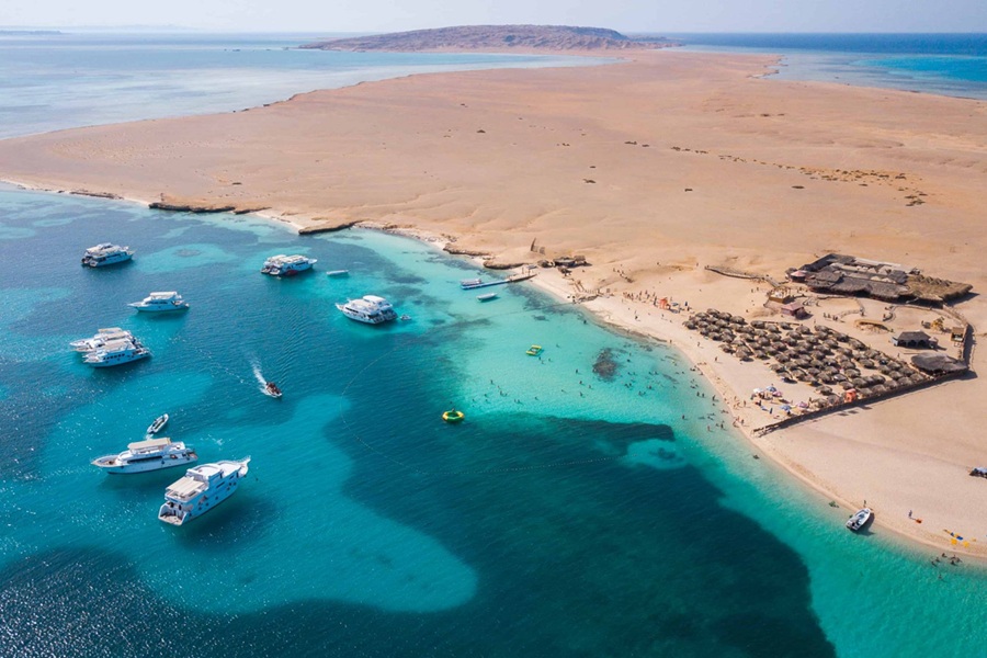 Varias embarcaciones navegan y fondean en aguas turquesas junto a una costa desértica con sombrillas y bañistas, ofreciendo una vista aérea de la isla en Hurghada, Mar Rojo.
