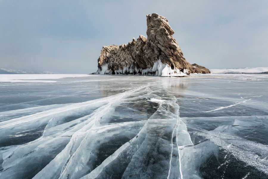 Lago Baikal: Vista panorámica de hielo resquebrajado sobre la superficie congelada del lago Baikal con una formación rocosa al fondo en Rusia.
