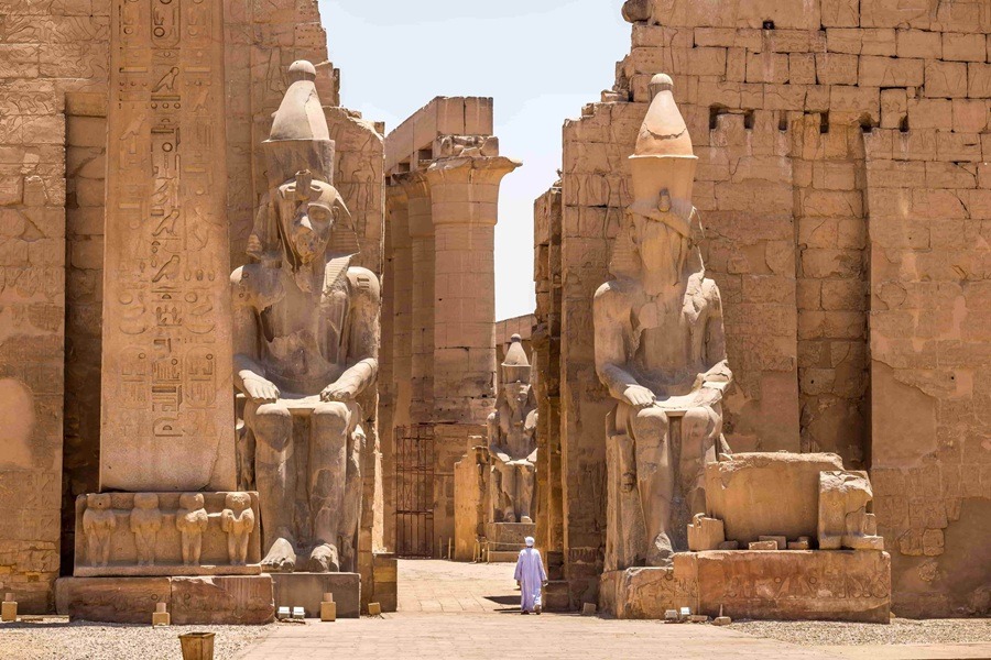 Dos colosales estatuas de piedra flanquean la entrada monumental del Templo de Luxor, mientras una persona con túnica blanca camina entre ellas hacia las columnas interiores.
