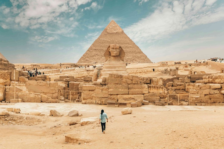 Una persona camina de espaldas por la arena dirigiéndose hacia la Gran Esfinge y la Pirámide de Kefrén en el complejo arqueológico de Guiza, Egipto, bajo un cielo parcialmente nublado.