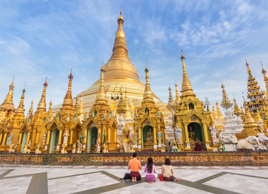Tres personas se arrodillan a orar frente a una monumental pagoda dorada rodeada de agujas e intrincados santuarios en un gran patio bajo un cielo azul.