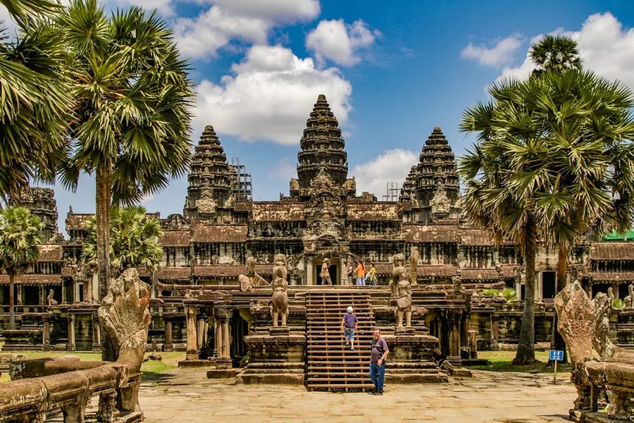 El templo de Angkor Wat se alza con sus imponentes torres de piedra mientras varios turistas recorren sus escalinatas, rodeados de palmeras bajo un cielo soleado.