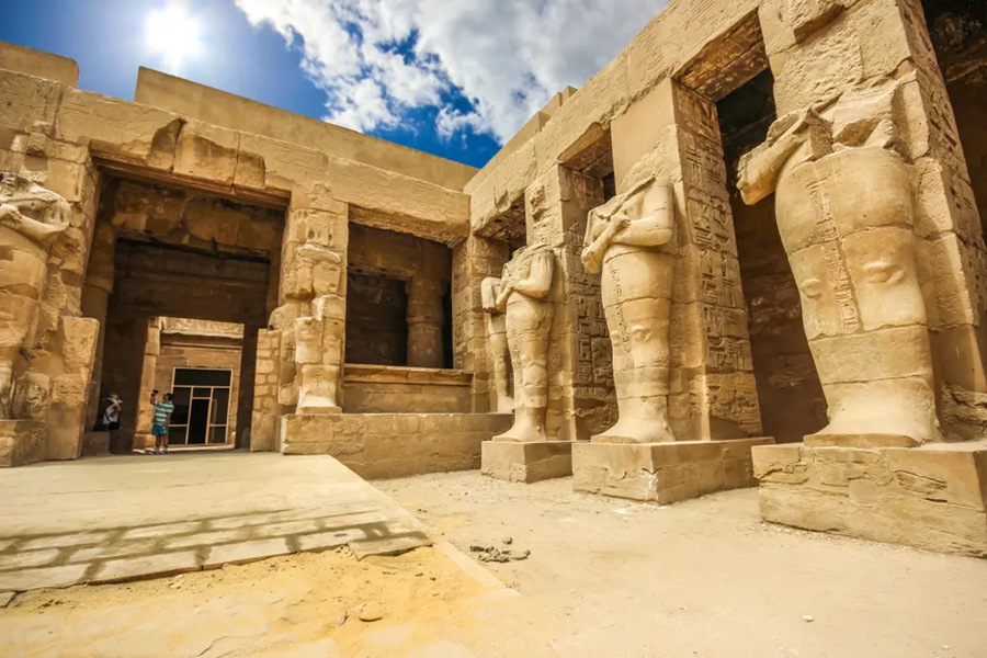 Grandes estatuas de piedra sin cabeza flanquean las columnas de un antiguo templo egipcio, iluminadas por un sol radiante bajo un cielo azul con nubes dispersas.