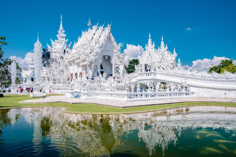 El intrincado Templo Blanco de Chiang Rai resplandece al sol, reflejándose sobre un estanque ajardinado con peces bajo un cielo azul despejado.