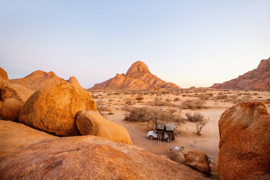 Un vehículo de safari acampando entre grandes rocas anaranjadas y vegetación dispersa en un desierto de montaña.