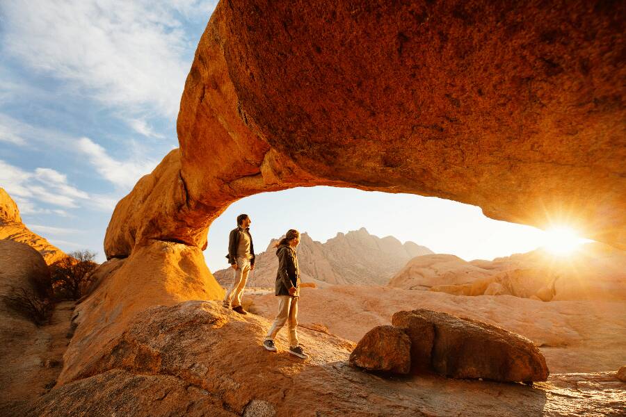 Dos personas contemplan el paisaje desde una formación rocosa en arco mientras la luz del sol se filtra entre las rocas.