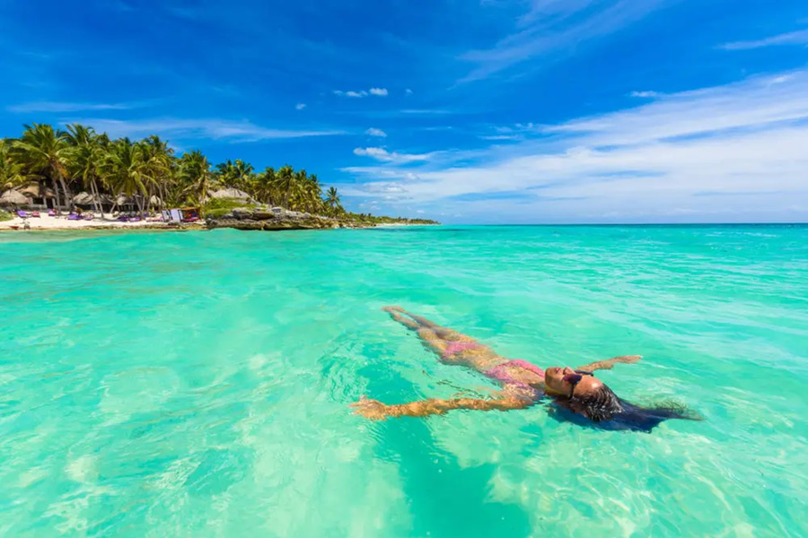 Una mujer en bikini flota relajada boca arriba sobre aguas turquesas y cristalinas, frente a una costa tropical repleta de palmeras bajo un cielo despejado.