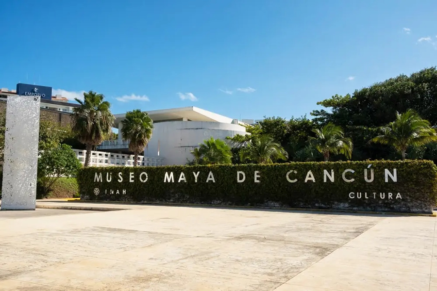 Un seto de entrada exhibe el letrero "MUSEO MAYA DE CANCÚN INAH CULTURA" sobre una explanada amplia, frente a un edificio blanco rodeado de vegetación tropical bajo un cielo despejado.