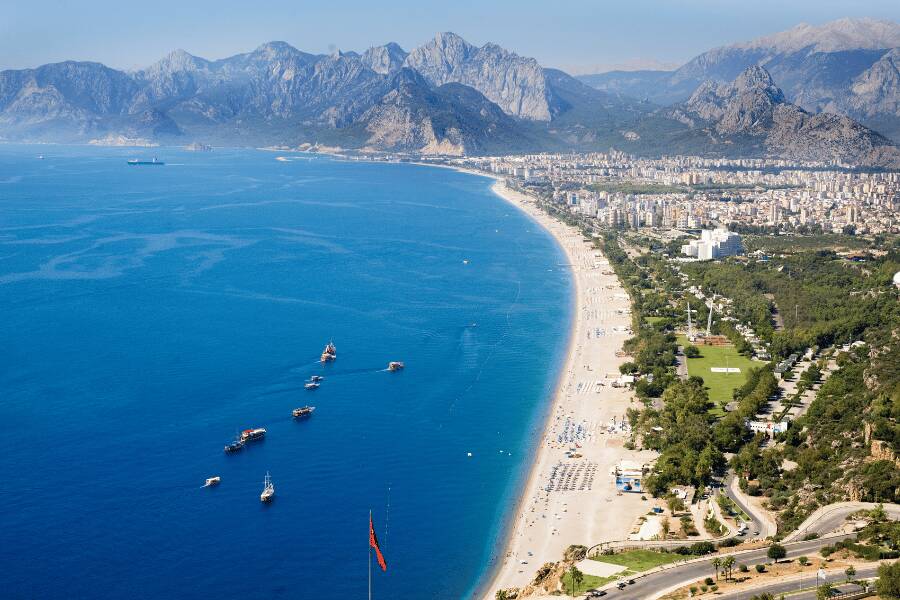 Una costa curva bordea aguas turquesas hacia una ciudad y montañas lejanas bajo un cielo despejado. Antalya