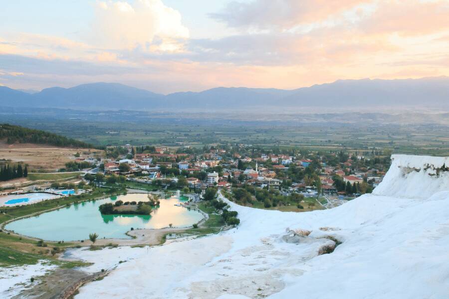 Terrazas de travertino blanco brillante descienden hacia un valle con un lago verde y colinas de fondo.
