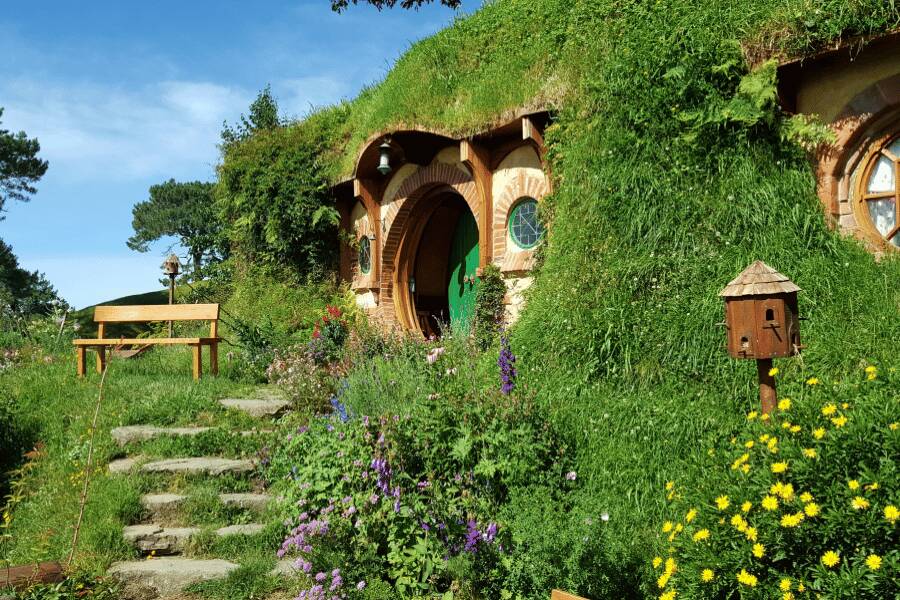 Una casa hobbit con puerta redonda y fachada de madera se integra en una colina verde llena de flores.