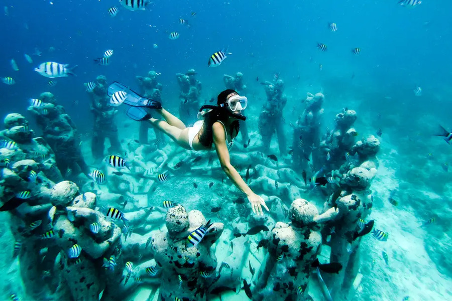 Una mujer nada haciendo snorkel sobre esculturas submarinas con forma humana, rodeada de peces a rayas en aguas cristalinas de color azul turquesa.