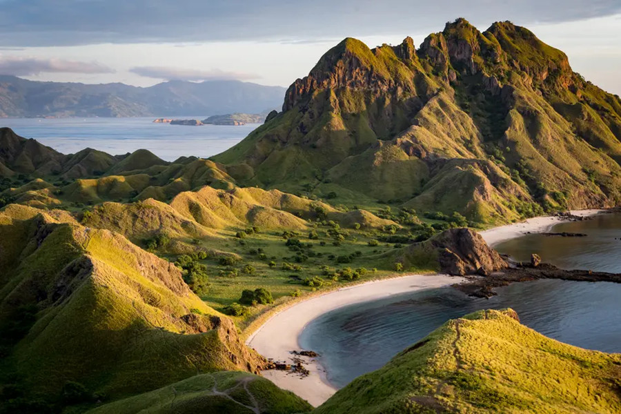 Montañas verdes y escarpadas se elevan junto a una bahía de arena blanca que se adentra en el mar calmo bajo la luz suave del atardecer.
