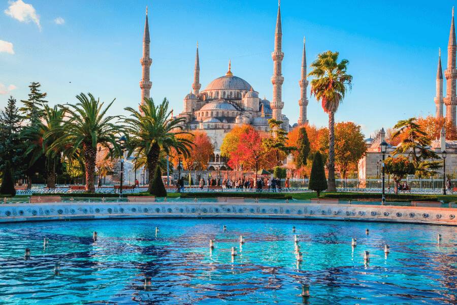 La Mezquita Azul en Estambul, Turquía, con sus seis minaretes, se ve detrás de una fuente de agua turquesa, palmeras y árboles con follaje otoñal bajo un cielo azul.